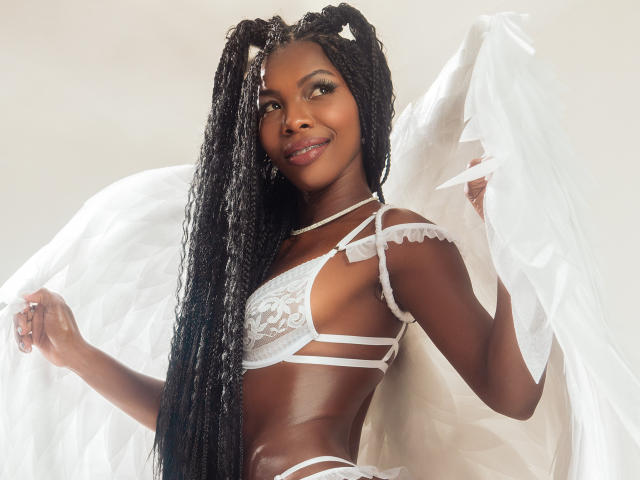 MichelleFlores with dark skin, long black braids, wearing white lace lingerie and angel wings, smiling, holding wings, showing her small tits and slim, toned ass.