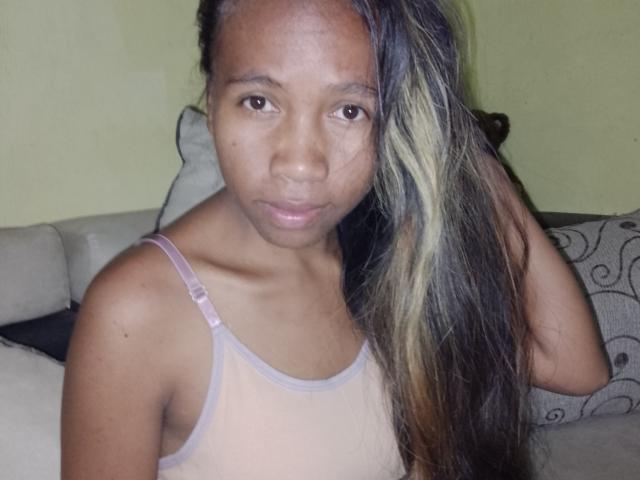 Ashra with long black and blonde hair, brown skin, small tits in a sheer pink tank top, looking straight at the camera, hand in hair.
