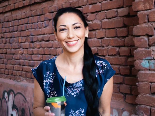 LiluGrants, smiling with dark hair in a ponytail, wearing a blue floral dress, holds a green-lidded cup with a straw, standing against a brick wall.