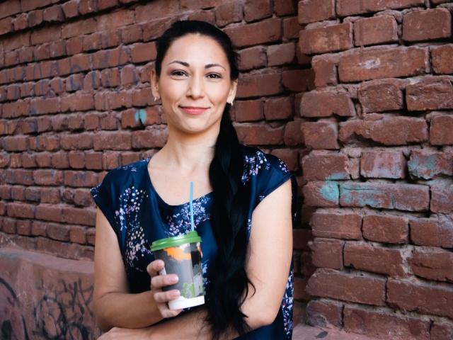 LiluGrants with long black hair, wearing a dark blue floral dress, holding a coffee cup with a green lid and blue straw, smiling at the camera.