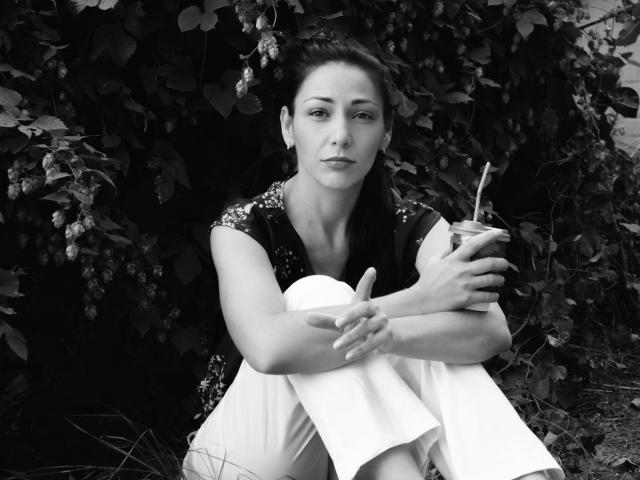 LiluGrants with dark hair, wearing a sequined top and white pants, sitting with knees up, holding a drink with a straw, in a black-and-white photo.