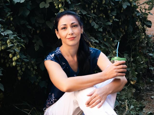 LiluGrants with dark hair, sitting, wearing a navy floral top and white pants, holding a green drink with a straw, smiling, arms crossed, looking cute and relaxed.