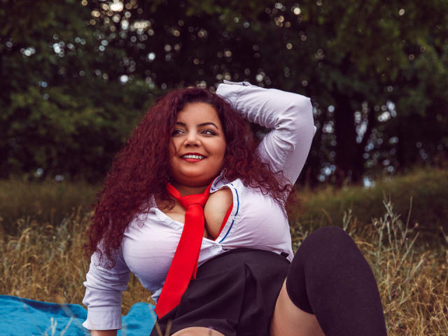 MiryamInes, avec ses cheveux roux bouclés, sa chemise blanche, son cravate rouge, sa jupe noire et ses bas montants, souriante, une main dans les cheveux, assise sur une couverture bleue, dans un champ herbeux, avec de grandes seins..