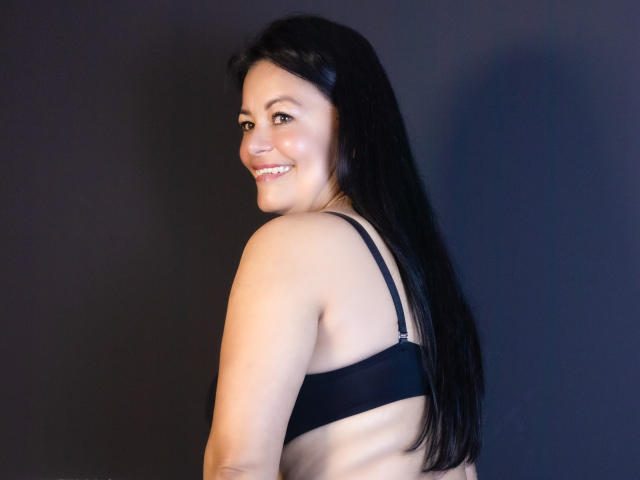 AnaMaturee, portant un soutien-gorge noir, souriant à la caméra, avec des seins de taille moyenne, de longs cheveux noirs, une peau claire, un maquillage discret, un fond sombre, une lumière douce, une pose légèrement de profil, attrayante..