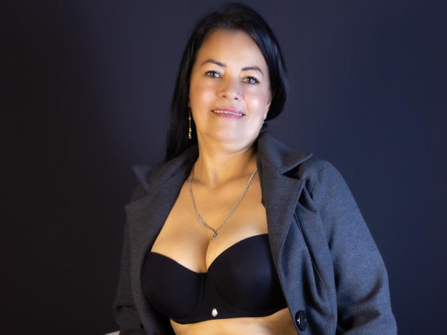 AnaMaturee with black bra, showing her tits, wearing a grey blazer, smiling, dark hair, earrings, necklace, against a black background.