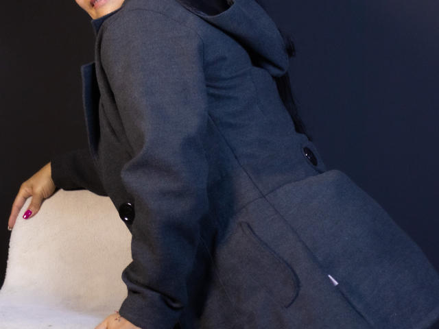 AnaMaturee in a dark gray hoodie, leaning on a white surface, butt facing camera, black buttons, pink nails, partial face visible, dark background.