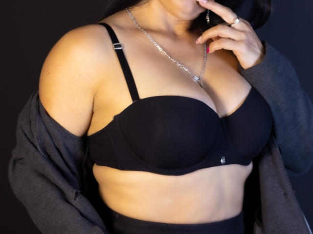 AnaMaturee's black bra, showcasing her big tits, with a silver necklace, dark cardigan off her shoulders, and her finger touching her lips. She has red nail polish.