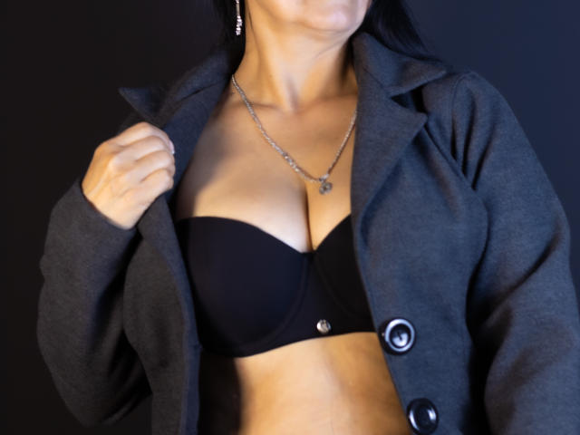Le soutien-gorge noir d'AnaMaturee révèle ses seins sous un manteau gris avec des boutons noirs, elle tient le manteau ouvert d'une main, et porte un collier en argent, sur un fond sombre..