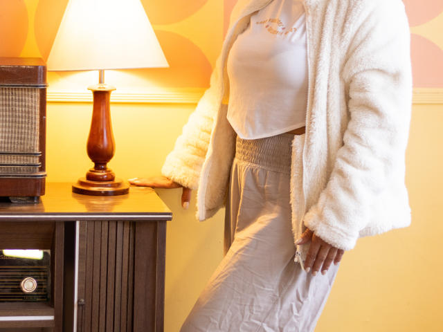 AnaMaturee with white fluffy jacket, white top, and high-waisted white skirt, standing next to wooden table with lamp and radio. Her tits are small and her ass is not visible.