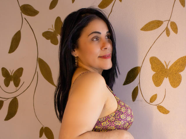 Girasol Walton, avec ses cheveux foncés, son rouge à lèvres rouge et son soutien-gorge violet à motifs, montrant ses seins, souriant à la caméra, avec une décoration murale en forme de papillon doré en arrière-plan..