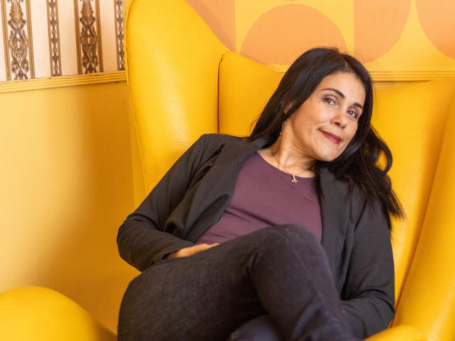 GirasolWalton, with black hair and a slight smile, sits in a yellow chair, wearing a black blazer, purple top, and black pants, arms crossed, showing a relaxed, confident posture.
