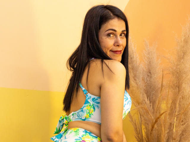 Girasol Walton, avec de longs cheveux noirs, regardant par-dessus son épaule, portant un bikini floral bleu et vert, souriant, montrant ses fesses, devant un mur jaune et orange, avec de l'herbe des pampas..