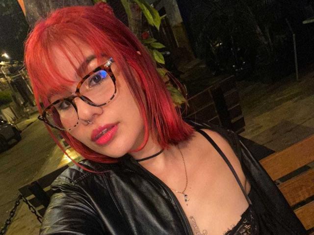 AriaSteele with red hair, glasses, nose ring, black leather jacket, and pink lipstick, showing her tits in a black lace top, sitting outside at night.