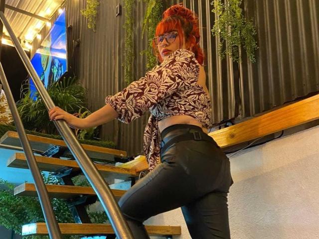 AriaSteele with red hair, glasses, and patterned top, showing her ass in black leather pants, climbing wooden stairs with a metal railing, holding a yellow bag.