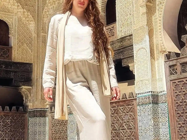 JazminBella-hot with long wavy brown hair, wearing a white dress and beige scarf, standing in a sunlit, ornately patterned building, showing her ass in white pants.