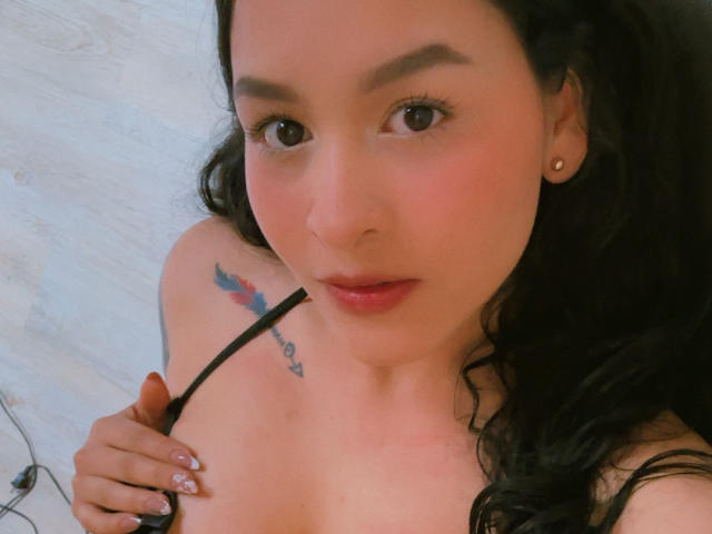 AyleenBloom with wavy black hair, light skin, and small earrings, showing a tattoo on her left shoulder, holding a black strap, and looking at the camera with a slight smile.
