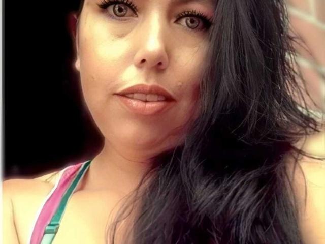 NatashaOlg has brown eyes, full lips, and black wavy hair covering one side of her face. She's wearing a pink and green top, looking at the camera.