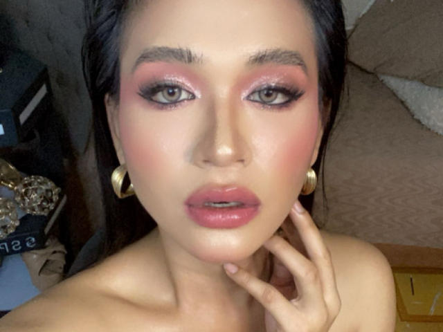 BettinaJohnson with pink eyeshadow, glossy pink lips, and dark makeup, wearing gold hoop earrings, touching her chin with her hand, looking into the camera with a slight pout.