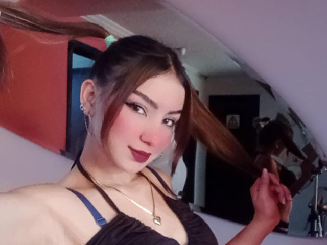 Ashley Miller, avec un blush rose, un rouge à lèvres foncé et un soutien-gorge noir, tenant ses longs cheveux bruns, portant un collier, dans un selfie pris devant un miroir. Seins visibles, mince, mignonne, d'origine asiatique, fond réfléchissant..