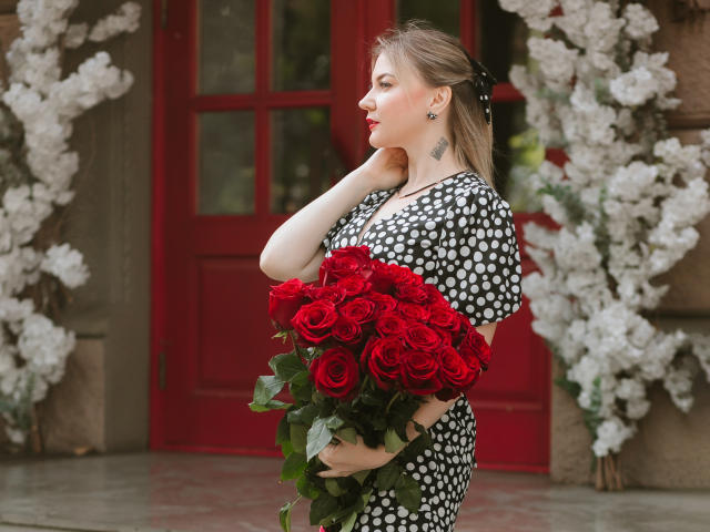 ParadiseRoxanne avec des roses rouges, une robe à pois, un nœud noir, tenant un bouquet, une porte rouge, des fleurs blanches, vue de profil, main sur le cou, regardant à droite, peau pâle, tatouage sur le côté gauche..