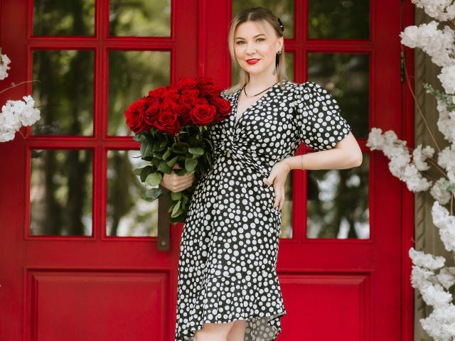 ParadiseRoxanne mit roten Rosen, in einem schwarz-weißen Punktekleid, vor einer roten Tür stehend, mit rotem Lippenstift lächelnd und den Blumenstrauß in ihrer rechten Hand haltend..
