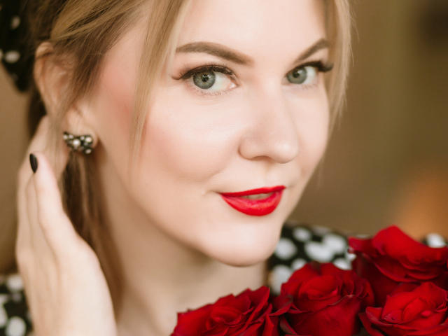 ParadiseRoxanne met rode rozen, rode lippenstift, zwarte nagellak en een zwart jurkje met stippen, die bloemen vasthoudt, de camera aankijkt met groene ogen en een lichte glimlach..