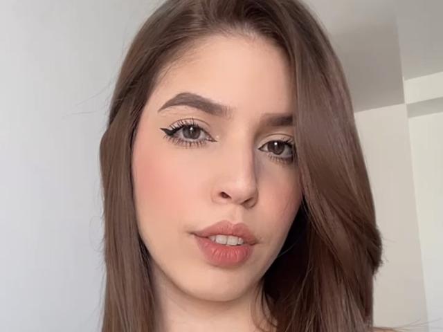 NatyBakers heeft bruin haar, volle, roze lippen en een lichte huidskleur met subtiele make-up, waaronder zwarte eyeliner. Ze kijkt iets naar rechts en heeft een neutrale uitdrukking..