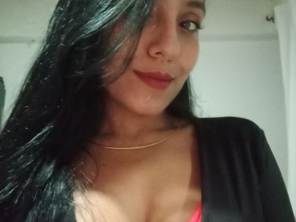 LuciBelly with long black hair, red lipstick, and a gold necklace, showing cleavage in a pink bra, wearing a black cardigan, smiling subtly.