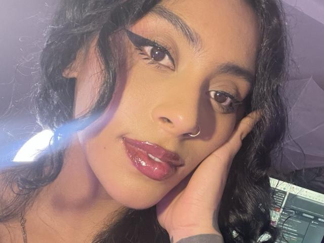 MagicMargo has glossy red lips, dark wavy hair, and wears a nose ring. She's touching her cheek with a hand, looking at the camera with sultry eyes.