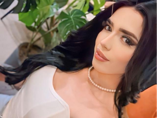 CatalinaPrincessX has long black hair, light skin, and is wearing a white top with a pearl necklace. She's smiling with brown eyes and full lips. Boobs are visible.