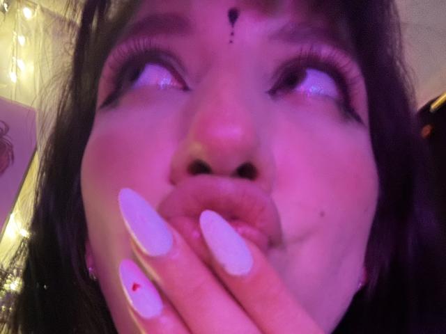 IzzysMisss's face with pink lighting, looking up, pouty lips, white nails with red dot, long eyelashes, nose, and a small beauty mark above her upper lip.