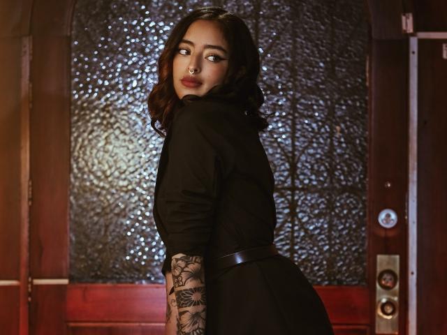 KathHoneyI with wavy dark hair, black dress, tattooed arm, and nose ring, looking over shoulder, dark, textured door behind. Boobs are covered, ass is in dress.