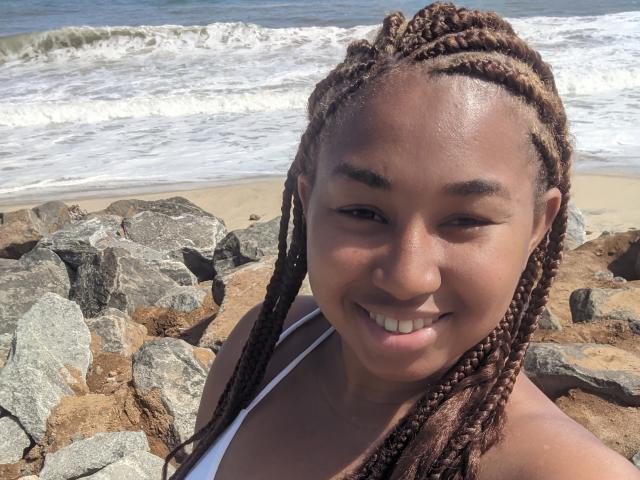 LoulouHot with brown braided hair, smiling at the camera, wearing a white bikini top, standing by rocky beach, waves in the background, showing off her cute ass.
