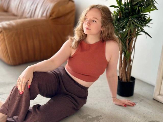SunnyBunny-hot with pale skin and blonde hair, wearing a rust-colored crop top and brown pants, sitting on a concrete floor with red nails, and a potted plant beside her.