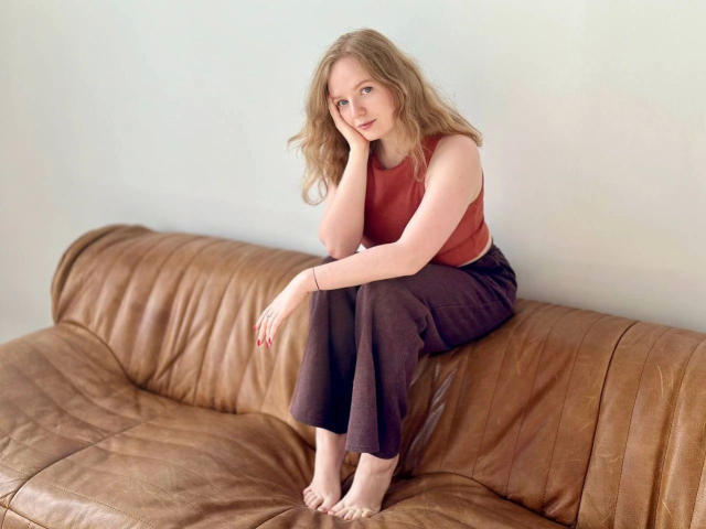 SunnyBunny-hot with wavy blonde hair, wearing a red tank top and brown pants, sitting barefoot on a brown leather couch, head resting on hand, looking at camera.