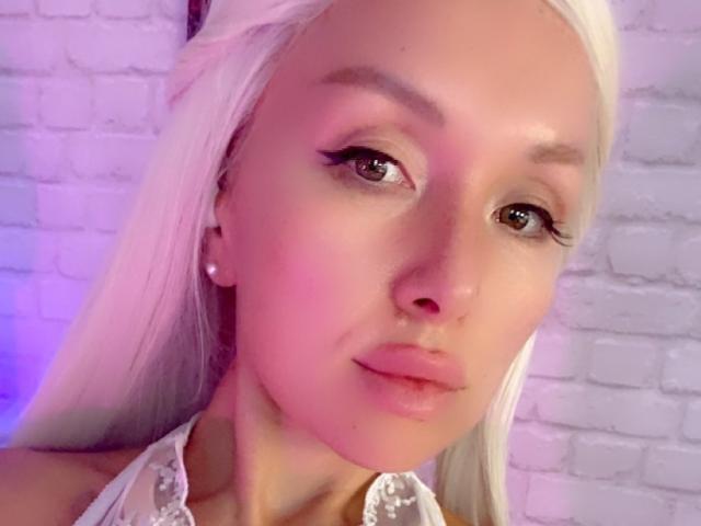 AvaGreenx mit blassrosa Haut, langem platinblondem Haar, braunen Augen und rosa Lidschatten. Sie trägt weiße Spitze, Perlenohrringe und hat einen neutralen Gesichtsausdruck..