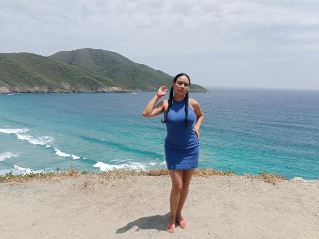 SamantaDark, com cabelo trançado, vestindo um vestido azul, em pé em um penhasco rochoso, segurando um pequeno objeto branco, olhando para a câmera, com um oceano azul e colinas verdes..
