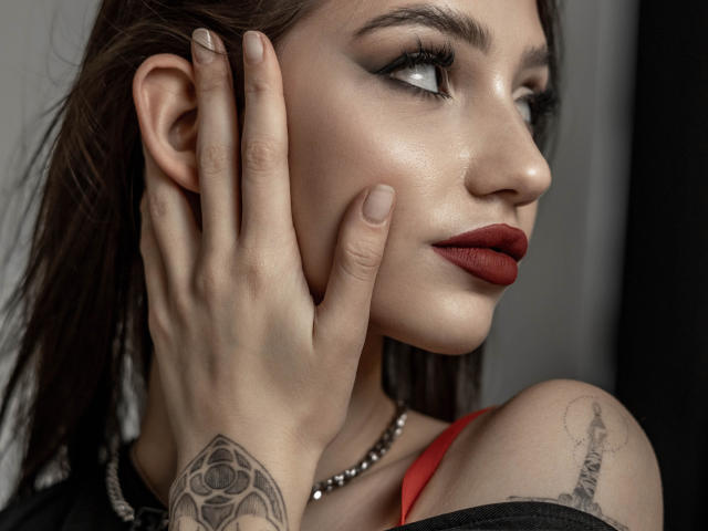 SophieKitty with her hand on her cheek, dark red lipstick, tattoos on her wrist and shoulder, black eyeliner, pearl necklace, and brown hair, looking to the side.