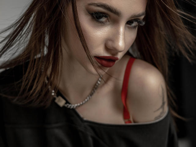 SophieKitty with dark brown hair, red lipstick, and red bra strap visible, wearing a black top and silver chain necklace, looking down with serious eyes. Boobs partially exposed.