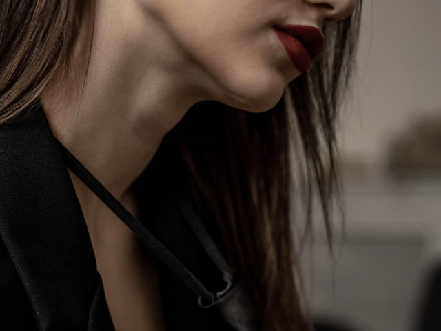 SophieKitty's face with dark red lipstick, slightly open mouth, black jacket, and lanyard, showing her neck and part of her chin, with brown hair in the foreground.