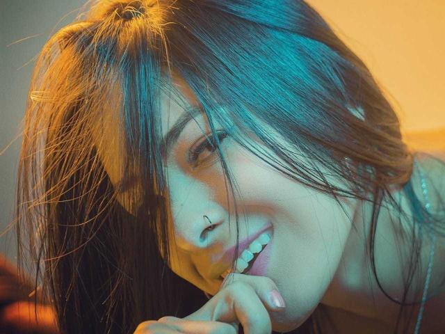 BellaHottieLove with messy brown hair, nose ring, and subtle smile, biting her finger, looking cute, has blue and yellow lighting, showing her sexy face and delicate features.