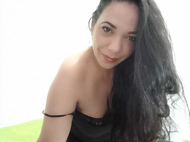 YolittS has long black hair, brown eyes, and a slight smile, wearing a black strap dress, showing her tits and a bit of her ass. She's looking at the camera.