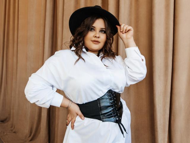 DreamForeYou with wavy brown hair, white shirt, black corset, and black hat, holding hat, standing, looking at camera, hand on hip, brown curtains in background.