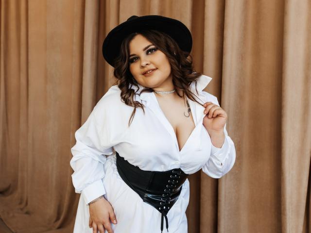 DreamForeYou with big tits, wearing a white button-up shirt, black corset, and black hat, standing in front of beige curtains, smiling, and touching her shirt.