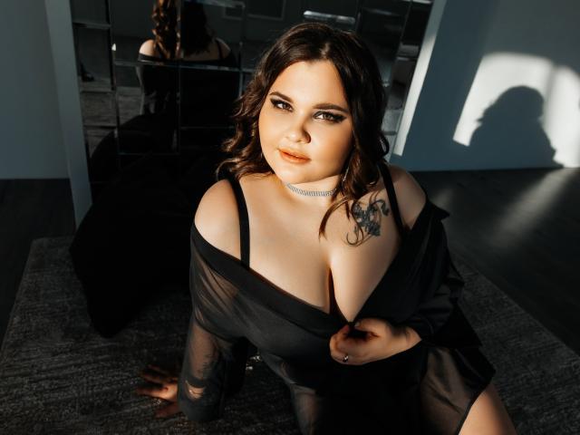 DreamForeYou has big tits, a tattoo on her right shoulder, and is wearing a black sheer top with her tits slightly exposed, sitting on a grey rug in sunlight.