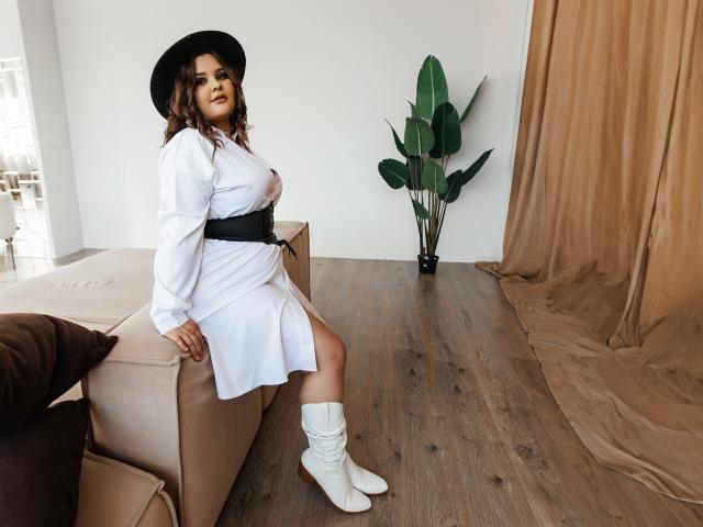 DreamForeYou in a white dress with black belt, white boots, and black hat, sitting on beige sofa, brown curtain, wooden floor, and green plant in background. Boobs visible.