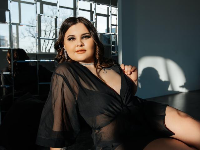 DreamForeYou with wavy brown hair, wearing a sheer black top, exposing her tits, sitting in sunlight, shadow on wall, choker necklace, looking at camera, hand on top.