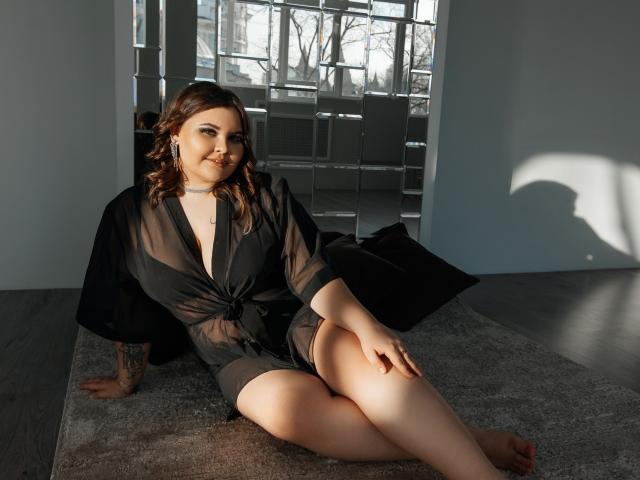 DreamForeYou with curly brown hair, wearing a sheer black robe, sitting on a gray rug, showing her tits and pussy, with a black pillow behind her, sunlight on her.