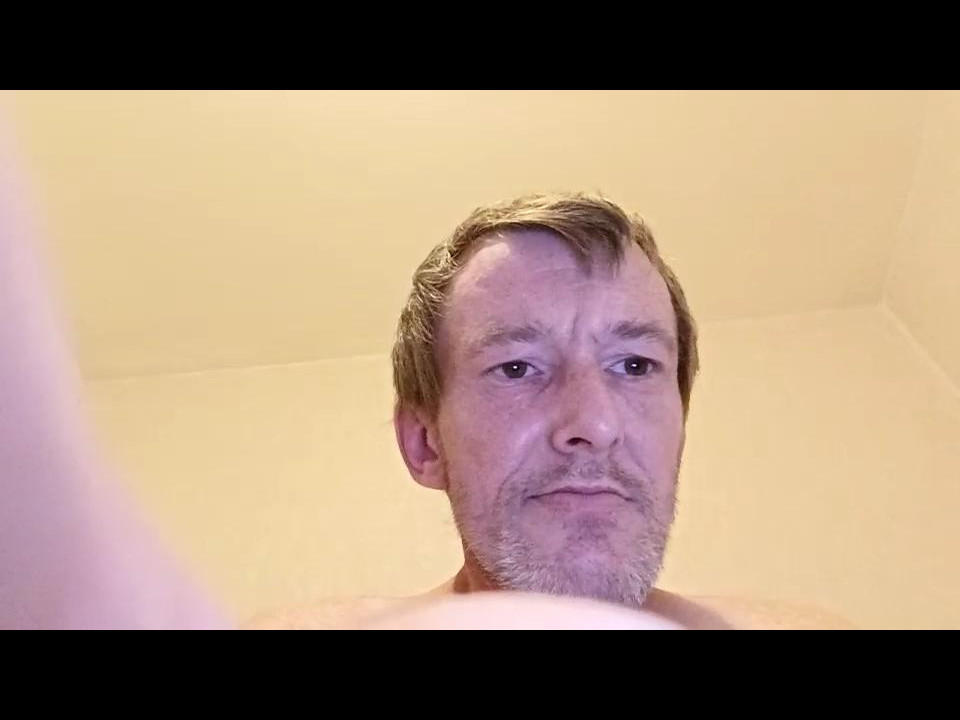 Hommcoquin with short brown hair and a light beard, shirtless, looking down with a slight smile, upper body in frame, from a low-angle perspective.