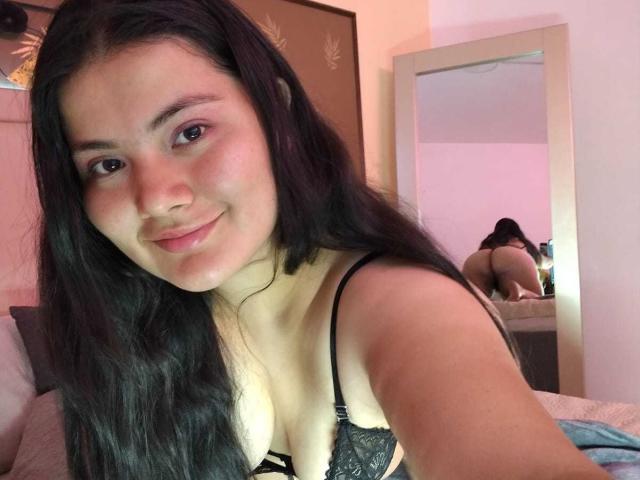 ZoeyClay with long black hair, pink eye shadow, and black lace bra, smiling at the camera. Her ass is visible in a mirror reflection, showing her in lingerie.